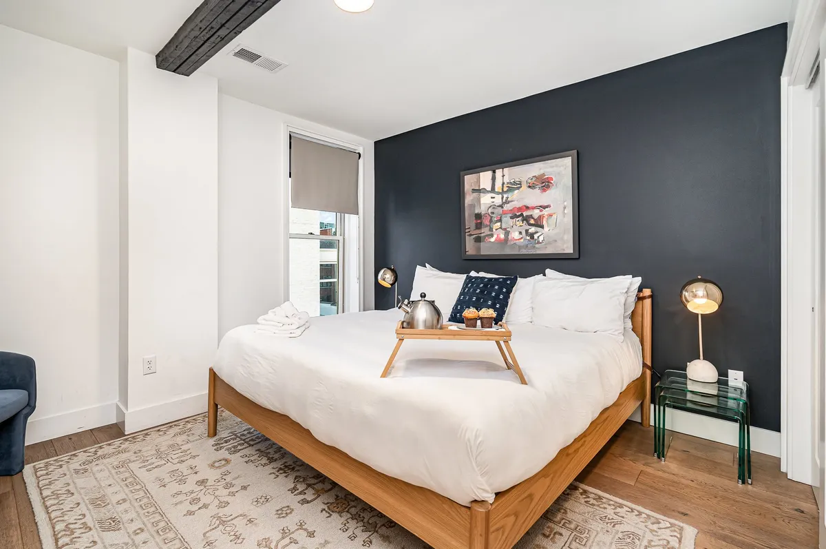 A serene bedroom featuring a plush bed with crisp white linens, modern artwork on the wall, and a tray with teapot and cups set atop the bedding. The atmosphere feels peaceful and inviting, perfect for relaxing after a long day.