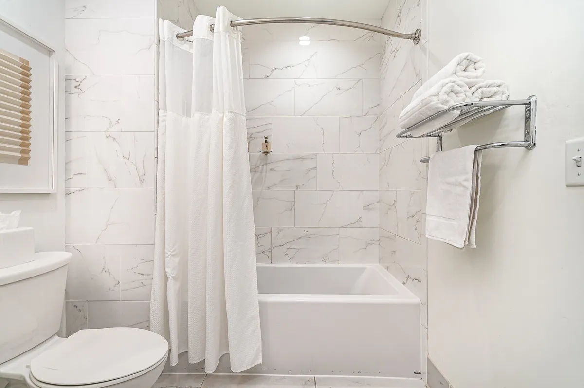 A spacious bathroom scene showcasing a bathtub with shower curtain, towel rack with fluffy towels, and a piece of framed wall art. The layout suggests a private space for relaxation and personal care.