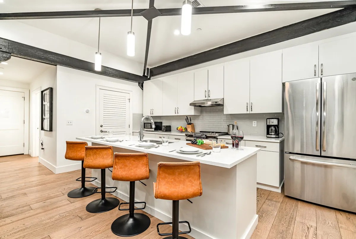 A bright, contemporary kitchen featuring a central island surrounded by bar stools, complete with high-end appliances, wine glasses, and food items on display. The space exudes a cheerful, welcoming atmosphere ideal for group gatherings, brunches, and casual entertaining.