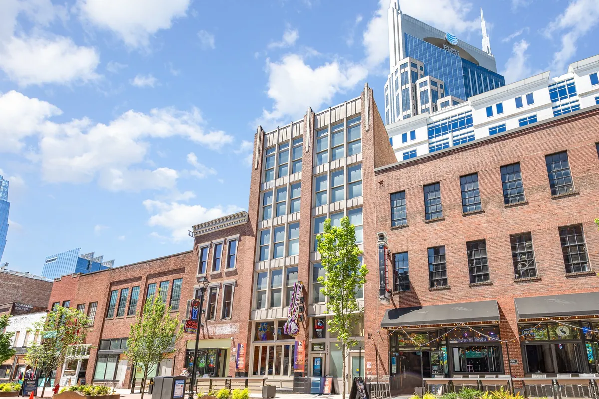 A vibrant downtown Nashville scene featuring a historic brick building juxtaposed with modern skyscrapers, leafy green trees, and a lively outdoor seating area. Groups can be seen dining al fresco and enjoying the energetic atmosphere of nearby shops and eateries.